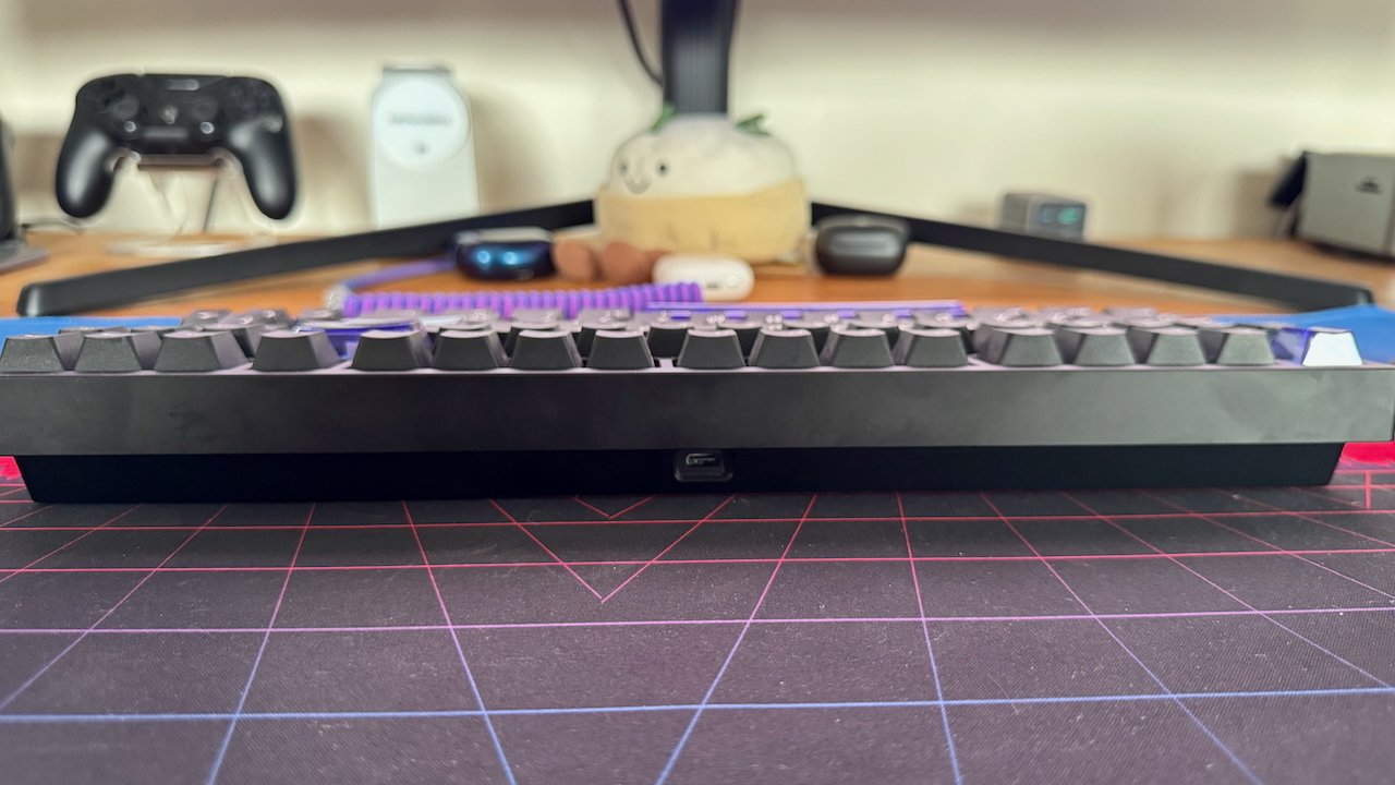 A low-profile mechanical keyboard on a grid-patterned desk mat, with various tech items and plush toy in the background.