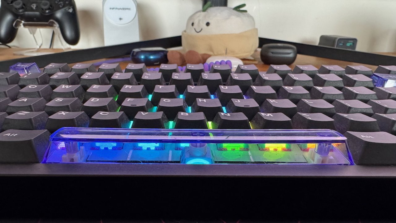 Close-up of a mechanical keyboard with colorful backlighting. In the background, a plush toy and gaming controller are visible on a desk.
