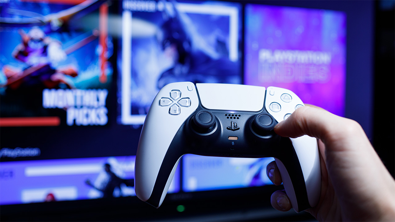 Person holding white PS5 controller, with blurred screen in the background displaying