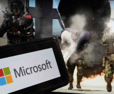 A smartphone displaying the Microsoft logo is held in front of a screen showing armed soldiers and an explosion in an action scene.