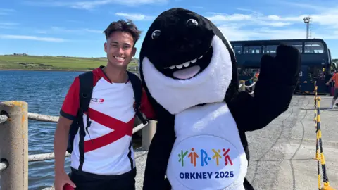 BBC Steven Mackay is stood at the harbour in Orkney smiling to take a photo next to the games mascot, Orla the Orca.