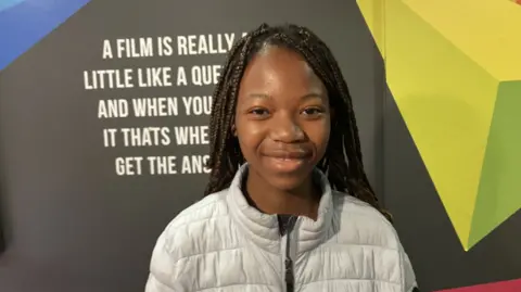 Shows a young black girl with breaded hair and wearing a grey zip-up puff jacket  smiling and standing in front a partially-eclipsed sign about film. 