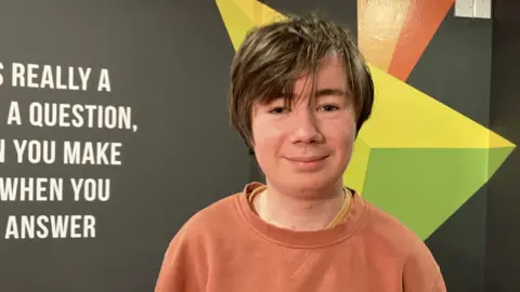 BBC Shows a teenage boy with hair parted from left to right, wearing an orange sweatshirt