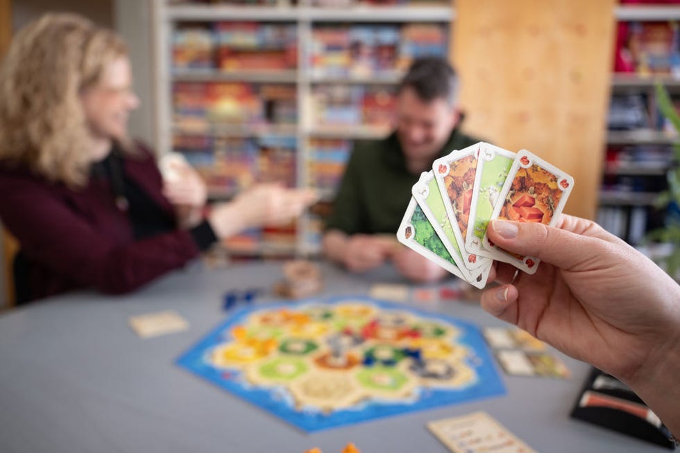 two people play the board game catan in the blurry background with a colorful game board on the table in front of them and someone holds a hand of game cards in the foreground two people play the board game catan in the blurry background with a colorful game board on the table in front of them and someone holds a hand of game cards in the foreground