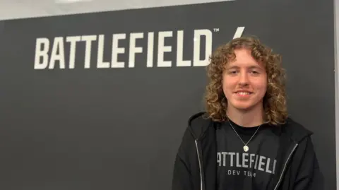 Black background with the words Battlefield 6 in white at the top of the picture. Vlad is wearing a black zipped hoodie with a black t-shirt underneath with Battlefield printed on the front. He has shoulder length curly light brown hair.