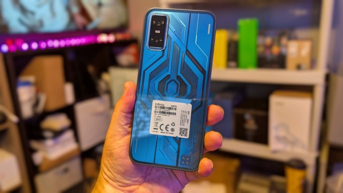 A person holding the Infinix GT 30 smartphone featuring a bold, cyberpunk-inspired blue back design with geometric etching and a label on the back, set against a blurred background of shelves with various electronic items.