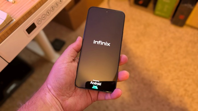 A close-up view of a person holding an Infinix smartphone displaying the startup screen with the brand logo 'Infinix' and 'Powered by Android' on a flat surface.
