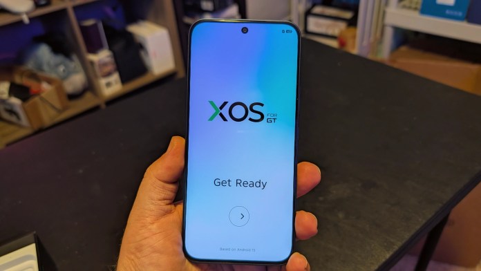 Infinix GT 30 smartphone displaying the XOS interface on its screen, held in a hand against a dark table backdrop.