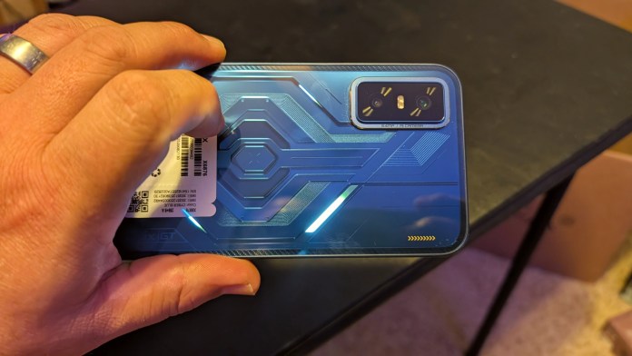 A close-up of the blue back panel of the Infinix GT 30 smartphone, featuring geometric designs and an illuminated LED pattern, held in a hand with a ring.