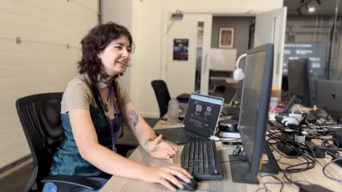James Vincent/BBC Yasmina Fadel from Maximalist sits at her desk at Production Park in South Kirkby. She is wearing a blue green dress and has a leaf tattoo on her left arm. She's got the computer mouse in her right hand.
