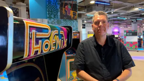 James Vincent/BBC Jamie Sefton from gaming industry body Game Republic wears a darks shirt with his hands clasped in front of him. He has grey hair and is smiling. He is standing in front of the Phoenix arcade machine at the National Video Games Museum in Sheffield
