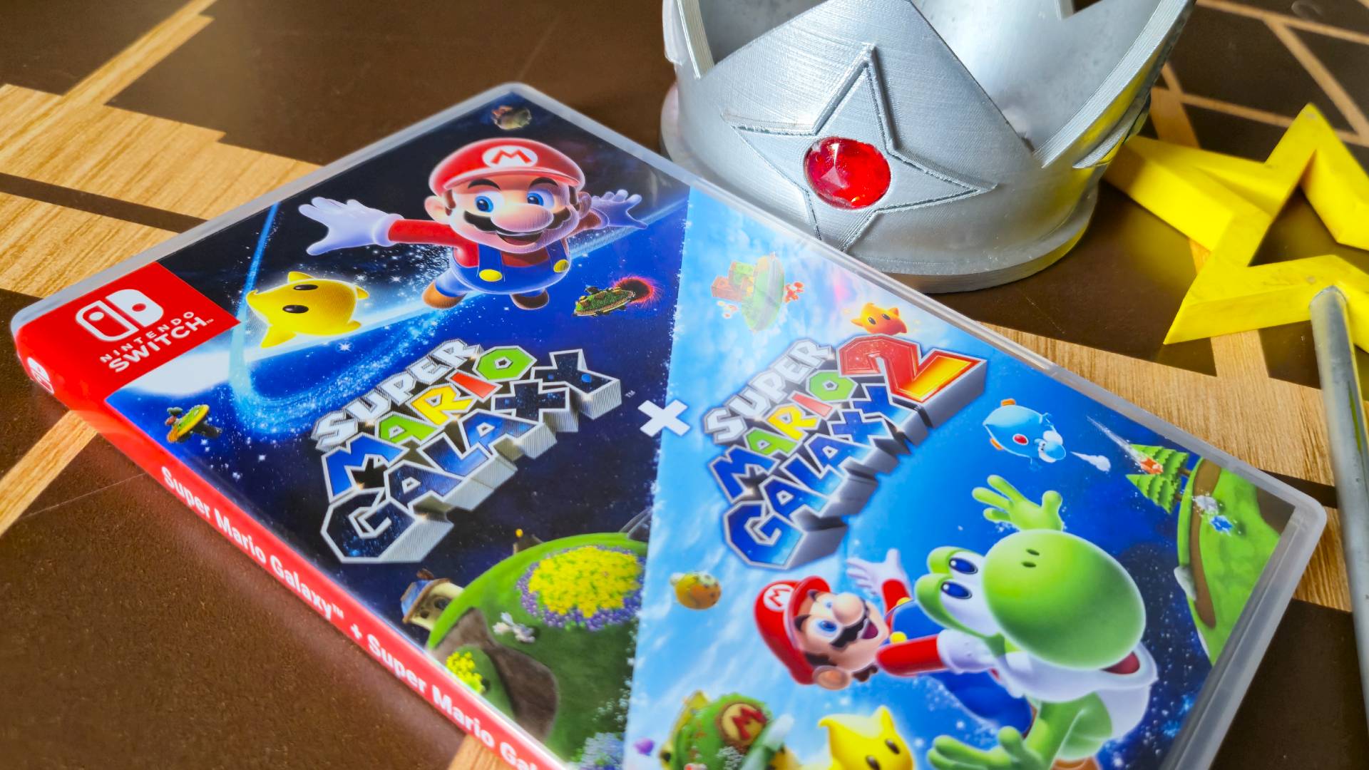 Close up of the front of the Super Mario Galaxy + Galaxy 2 Switch box, with a replica of Rosalina's crown and wand in the background.