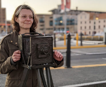 Game designer Susannah Emery stands behind a statue of a camera. She is a white woman with brown eyes and brunette hair. She is wearing glasses and a muted green jacket.
