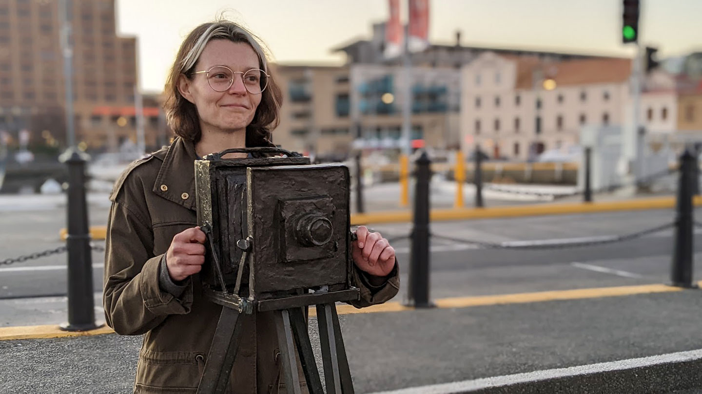 This game designer shares neurodivergent experiences through gaming Game designer Susannah Emery stands behind a statue of a camera. She is a white woman with brown eyes and brunette hair. She is wearing glasses and a muted green jacket.
