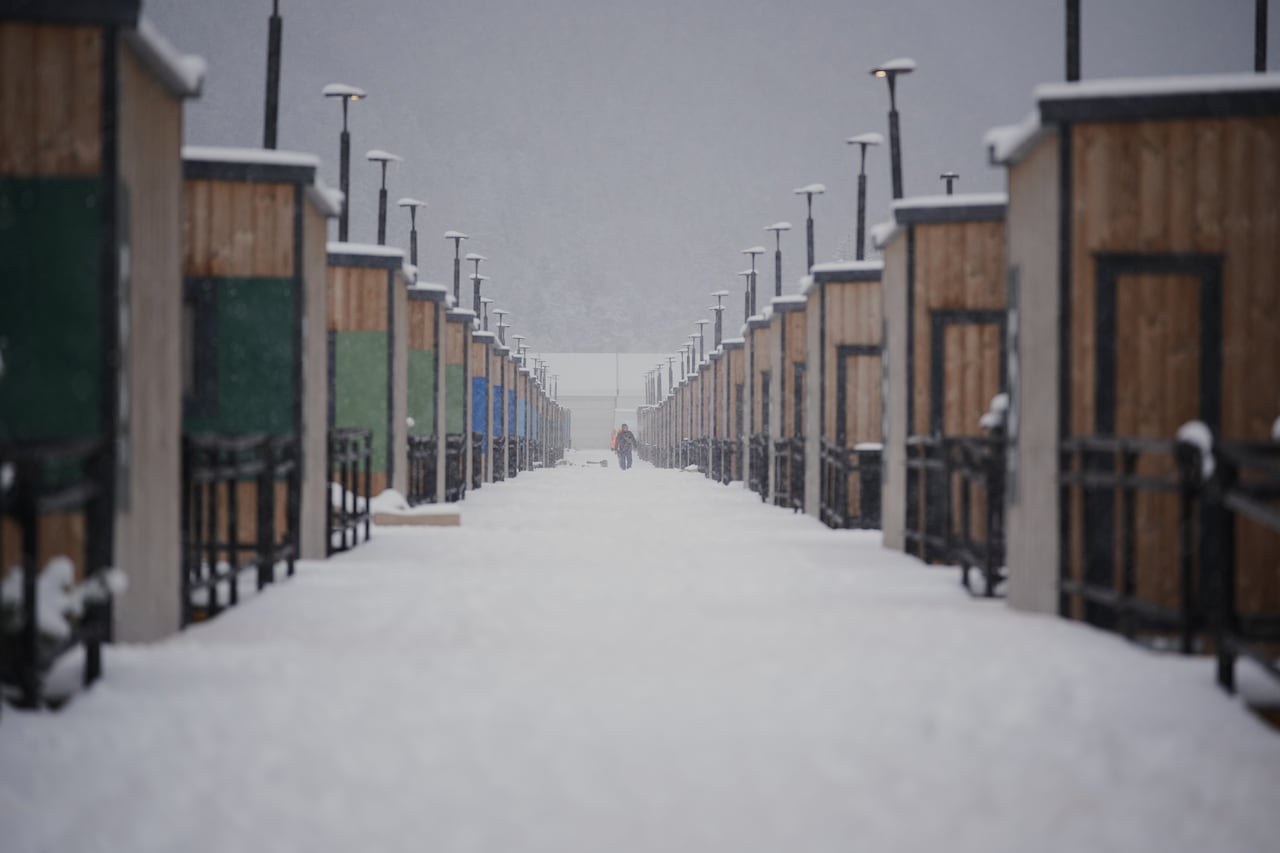 A person in winter gear walks down a snowy corridor between two sets of winter cabins covered in snow. 