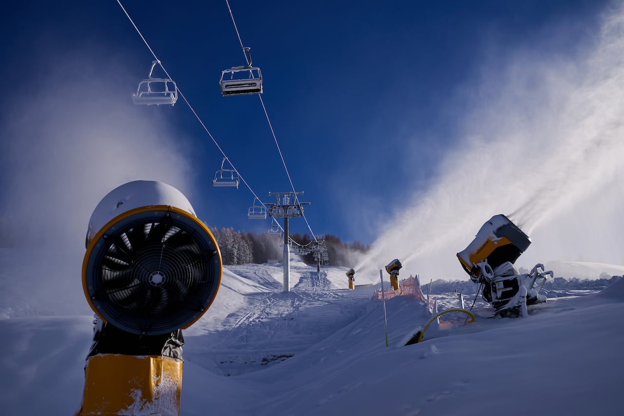 Four large yellow snow cannons blast clouds of snow onto a ski hill in various directions as a ski lift operates in the background. 