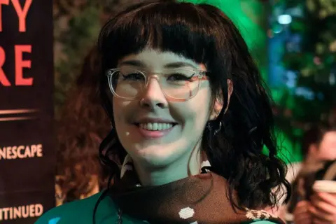 Molly Molly smiles at the camera. She is a woman with long black hair cut into a fringe and glasses. 