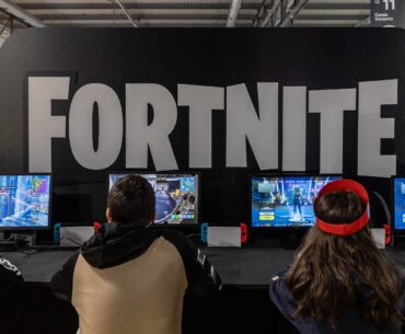 People playing "Fortnite" at a gaming event, seated at monitors with controllers. Large "FORTNITE" sign in the background.
