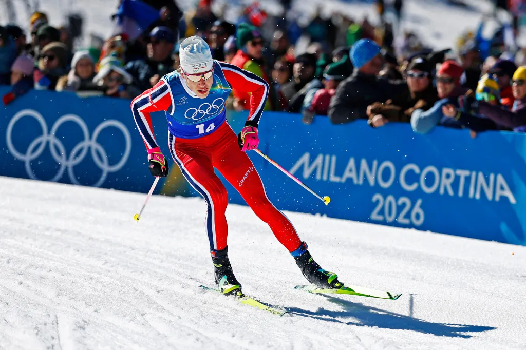 A cross-country skier races across snowy ground during a competition, with a sign reading “Milano Cortina 2026” behind.