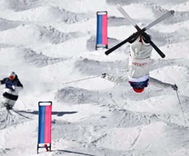 A freestyle skier flips upside down in midair over a snowy course during a competition.