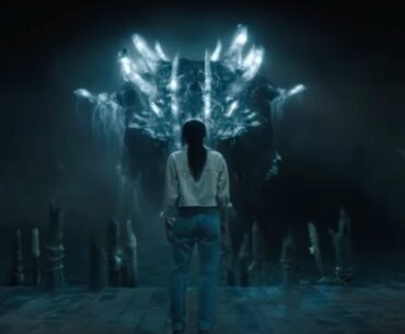 young woman in a dark cave standing with back to camera facing a gigantic monster glowing blue