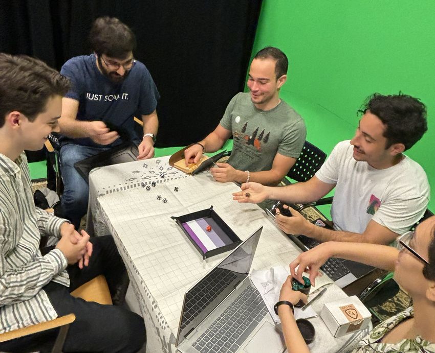 Five people sit around a rectangular table with a tray in the middle for rolling dice in the game Dungeons and Dragons, a TTRPG. One person has just rolled out a handful of dice, and another is holding one die over the table.