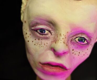 Clay sculpted pale face with prominent lips, large forehead, freckles.