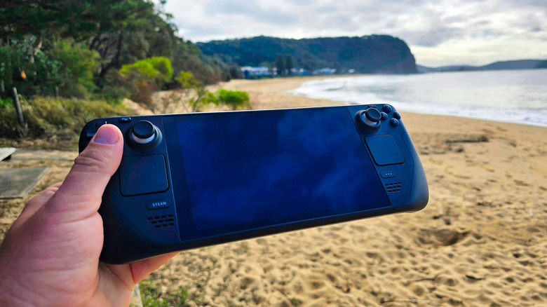 Hand holding a Steam Deck handheld gaming console with the beach and ocean in the background.