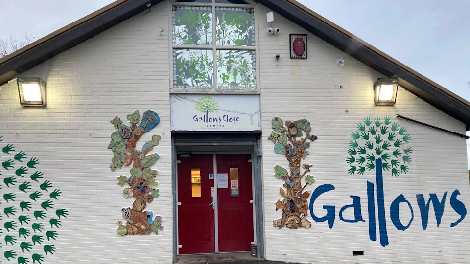 The front entrance of a single‑storey community-style building with a peaked roof. 

The exterior walls are painted white, and the entrance features two bright red double doors in the centre. Above the doors is a sign that reads “Gallows Close Centre”, accompanied by a tree-like logo.

On both sides of the doors are colourful, decorative murals of trees.