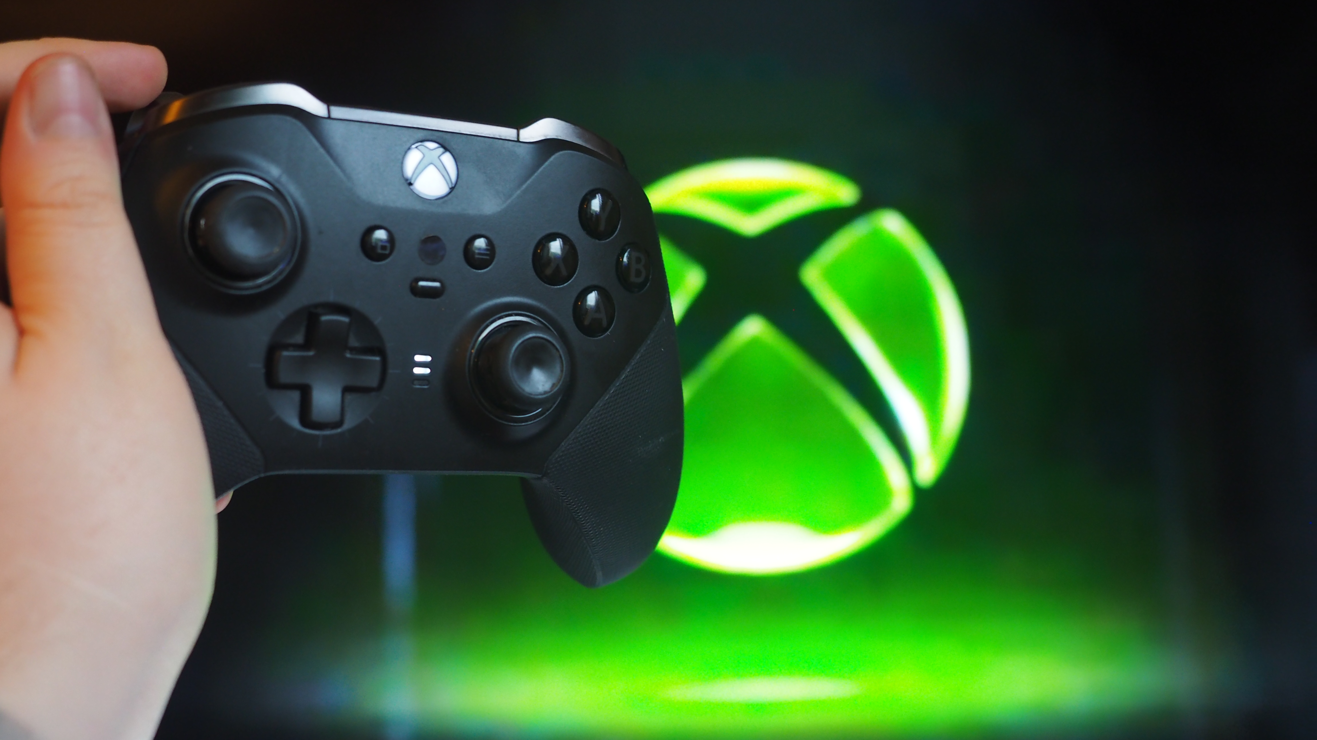 A hand holds a black Xbox controller against a glowing green Xbox logo on a screen. The scene conveys excitement and anticipation for gaming.