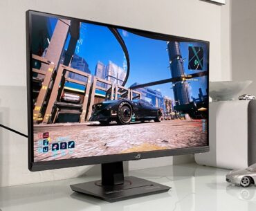 A sleek gaming monitor displays a vibrant cityscape from a video game. Next to it, a model car and speaker sit on a white desk, creating a modern tech vibe.