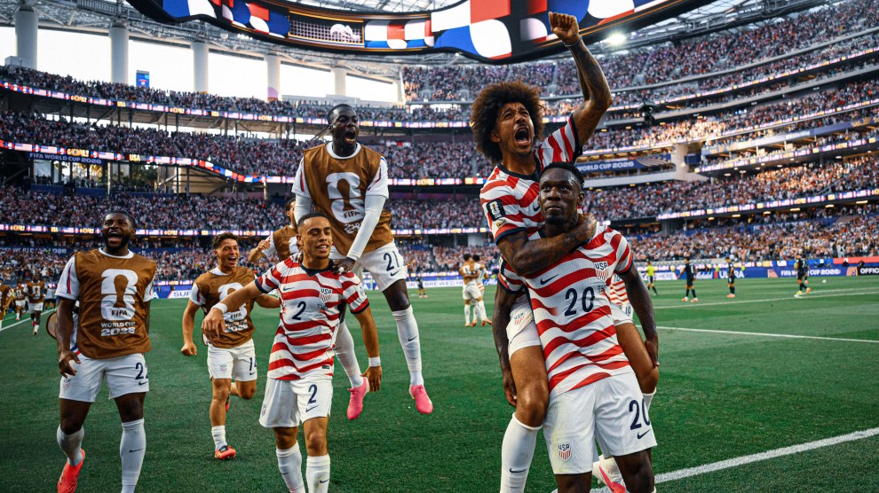 A Black soccer player stares defiantly at the crowd as a teammate hops onto his back with a celebratory raised fist. The US teammates are dressed in red-and-white striped shirts with white shorts. Behind them on the field, several team members rush to greet them in front of a stadium filled with fans.