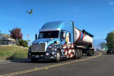 A virtual semi-truck with an American flag design on a highway, with a house, car, and airplane in the gameplay scene of American Truck Simulator.