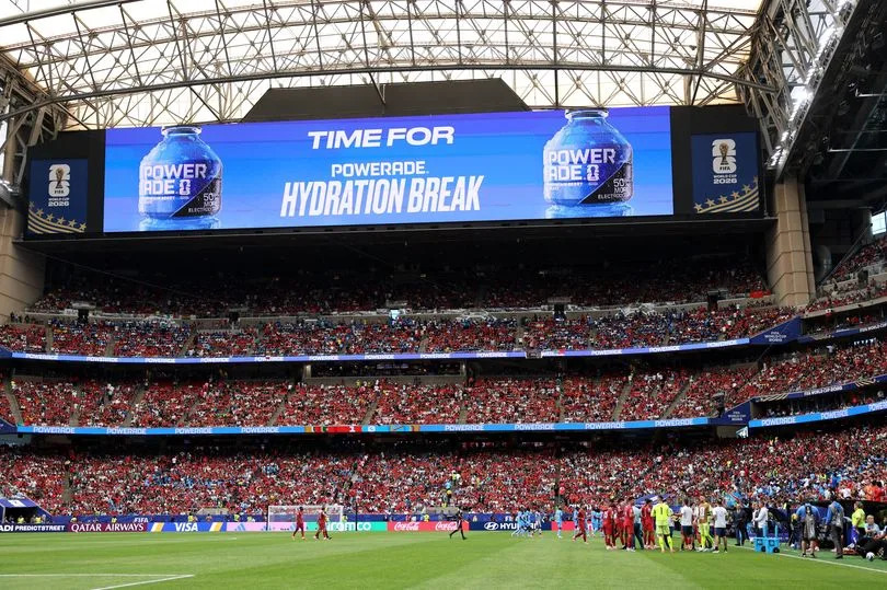 An indoor stadium filled with spectators during a sports event. A large digital screen above the stands displays a message, while the crowd and players are visible on the field below.