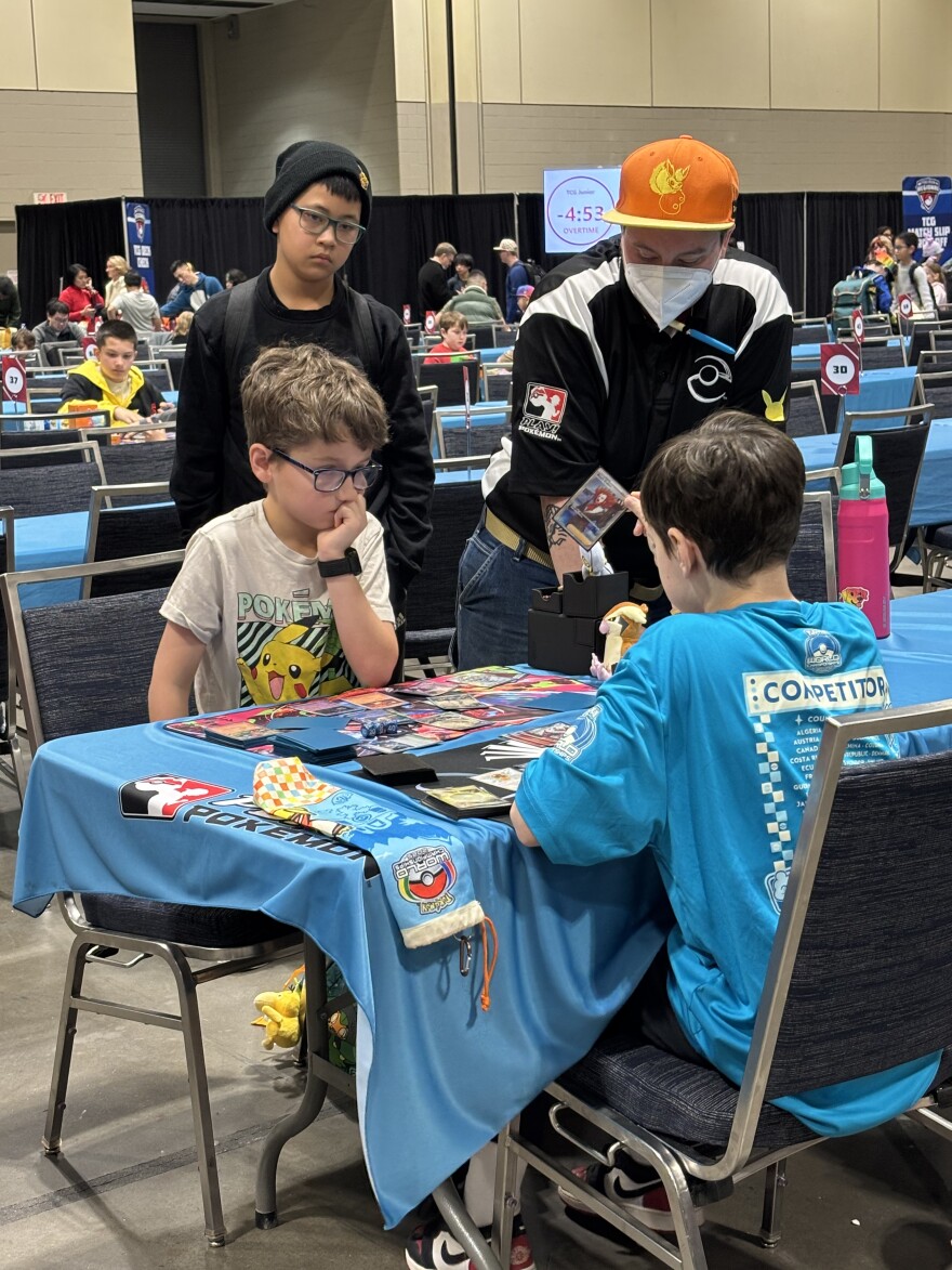 Two boys sit around a table. Another one stands beside them and an adult judge looks at the game they are playing