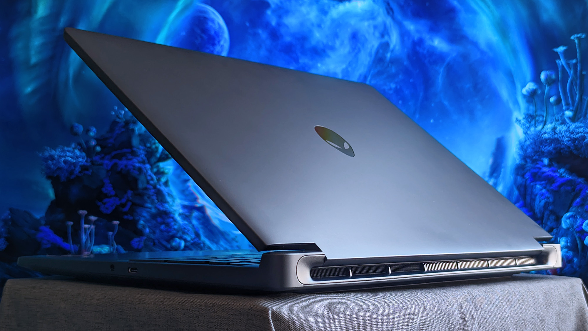 A partially open Alienware 15 laptop rests on fabric, showing its metallic lid with the Alienware logo and rear cooling vents, set against a glowing blue sci‑fi background.