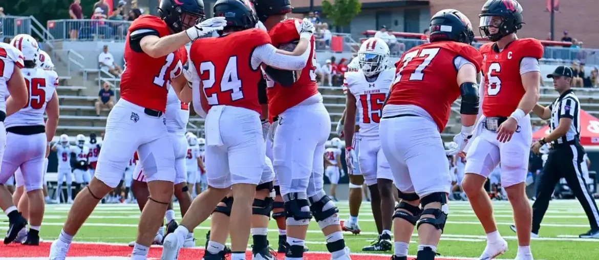 College football players in red uniforms huddle together on the field during a game, celebrating or planning a play at the line of scrimmage.